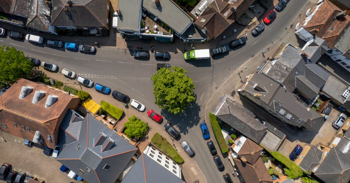 Aerial view of a UK town crossroads with terraced rooftops, parked cars and a large tree.